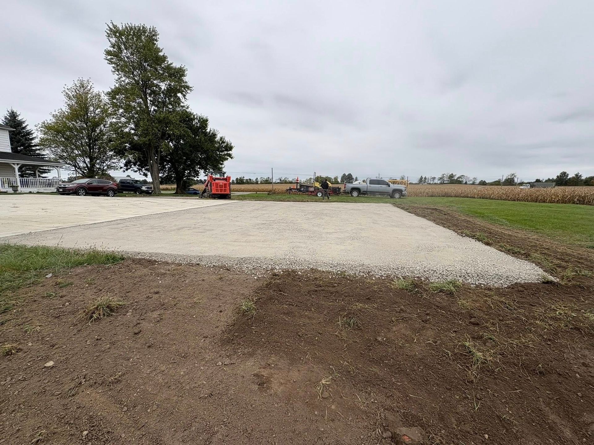 A gravel driveway pad in a rural setting, with cars and trees in the background under a cloudy sky.