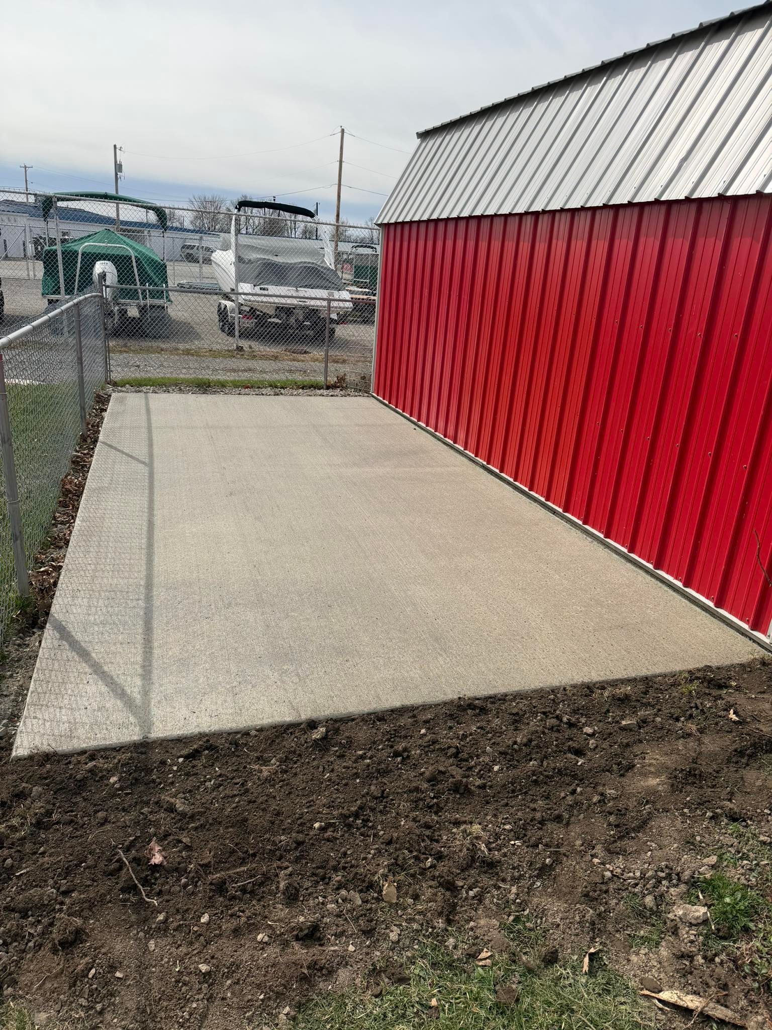A concrete patio area alongside a red, metal-sided building, with a chain-link fence and boats visible in the background.