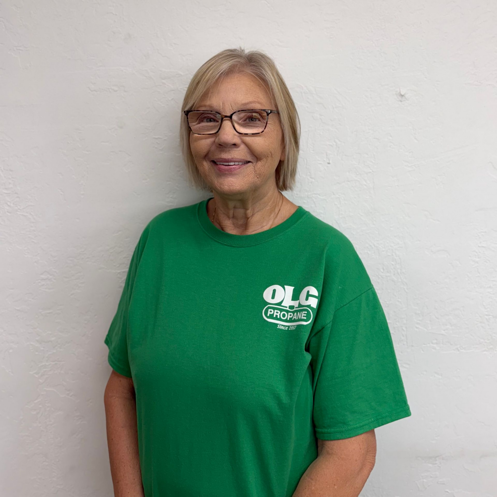 Woman in green shirt, glasses, smiling, standing against a white wall.