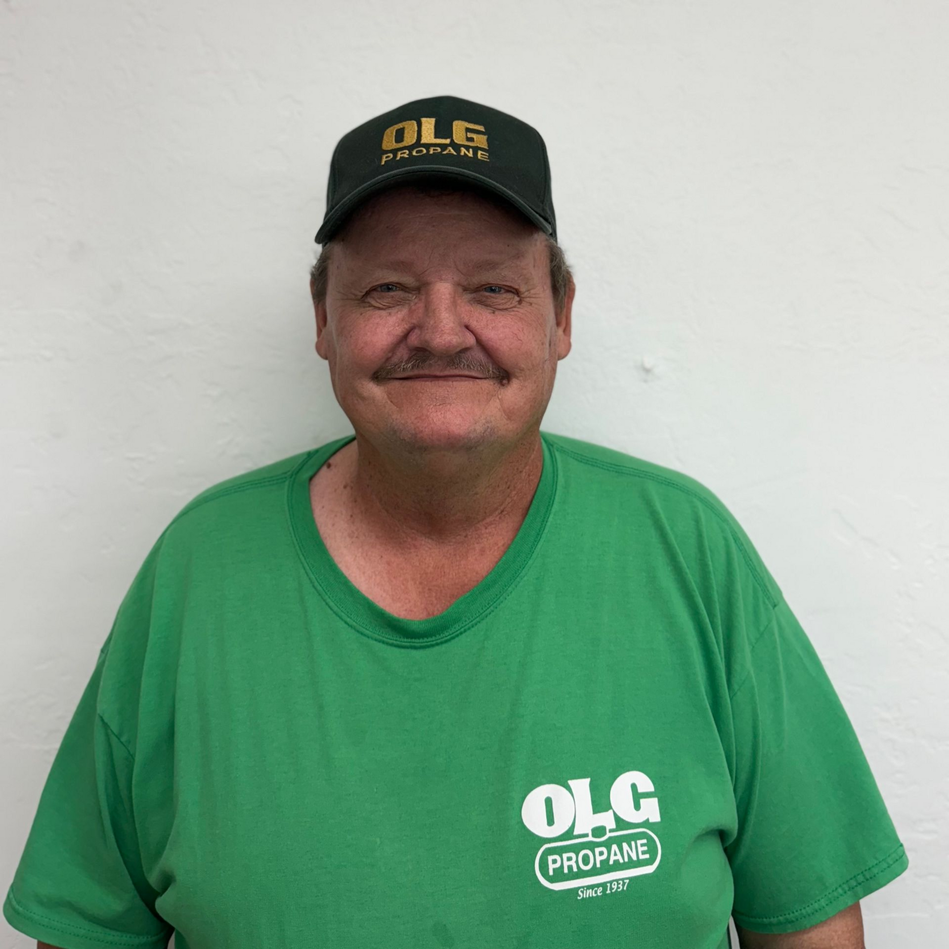 Man in green OLG Propane shirt and cap smiles against a white wall.