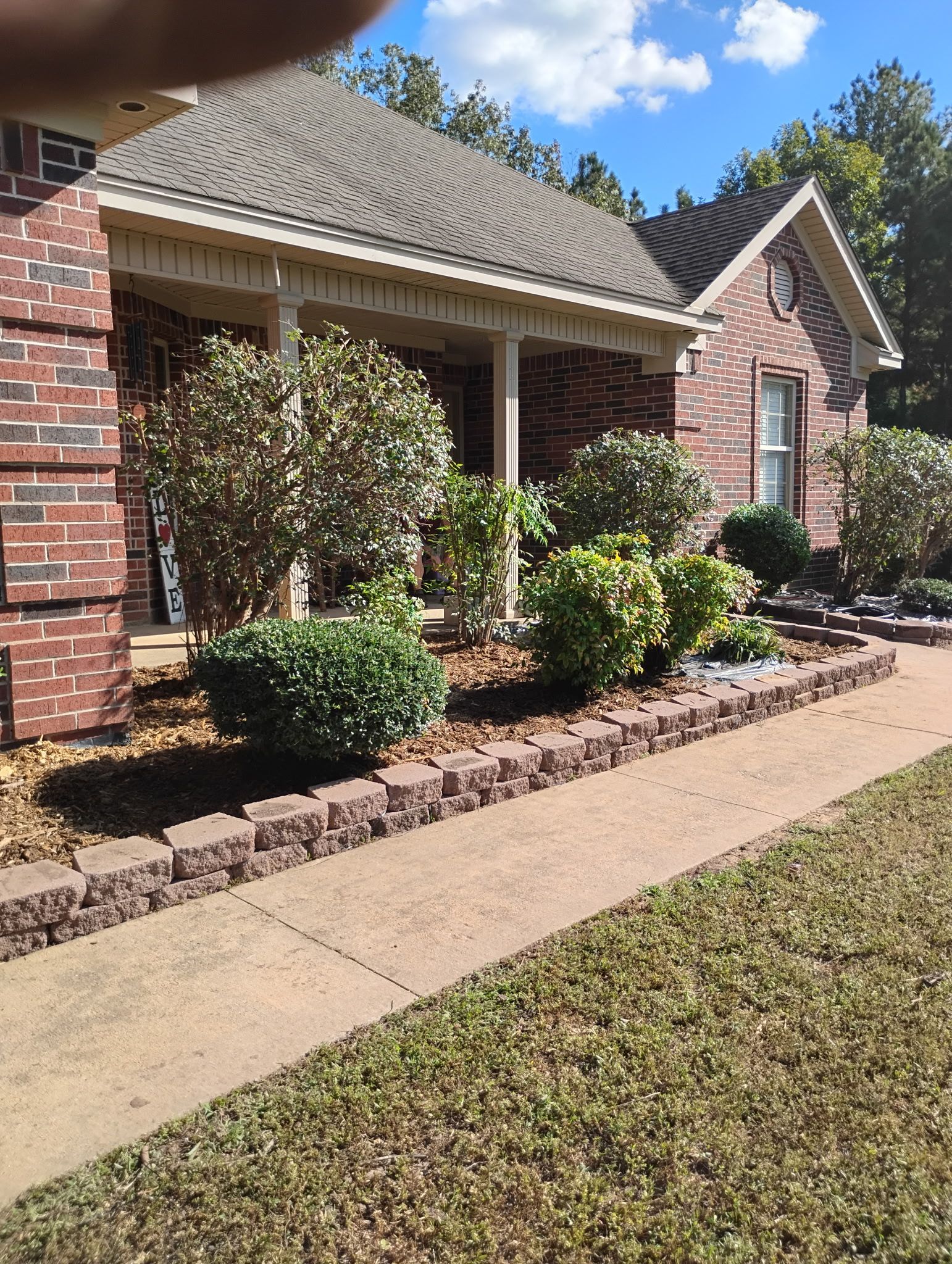 Red brick house with porch, flower bed bordered by dark bricks, walkway, blue sky.