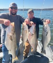 Two people smiling on a boat, each holding two large striped bass against a bright, sunny water background.