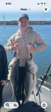 A person in a tan hoodie smiles while holding two large striped bass on a boat in the middle of a body of water.