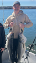 A person in a tan hoodie smiles while holding two large striped bass on a boat in the middle of a body of water.