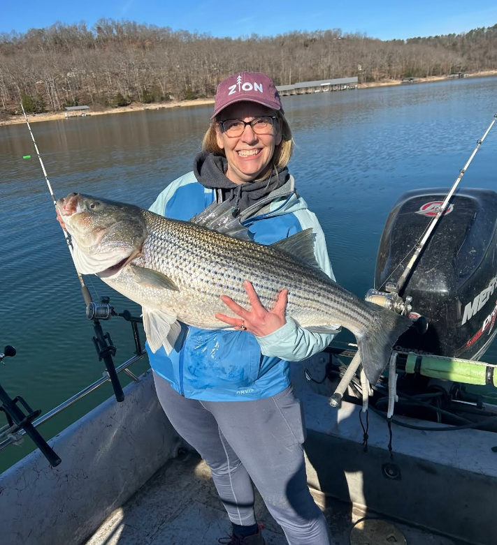 Woman on a boat holds a large striped bass fish, smiling. Blue water and a motor in background.