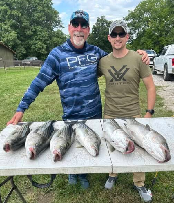 Two men pose with six large fish on a table outside. The older man smiles.