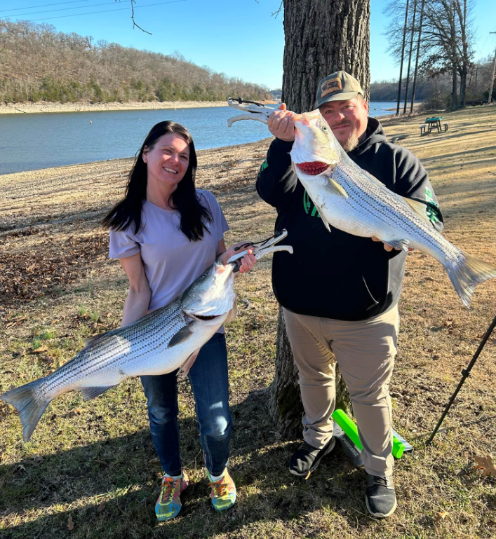 Woman and man smiling, holding large striped fish near a lake.
