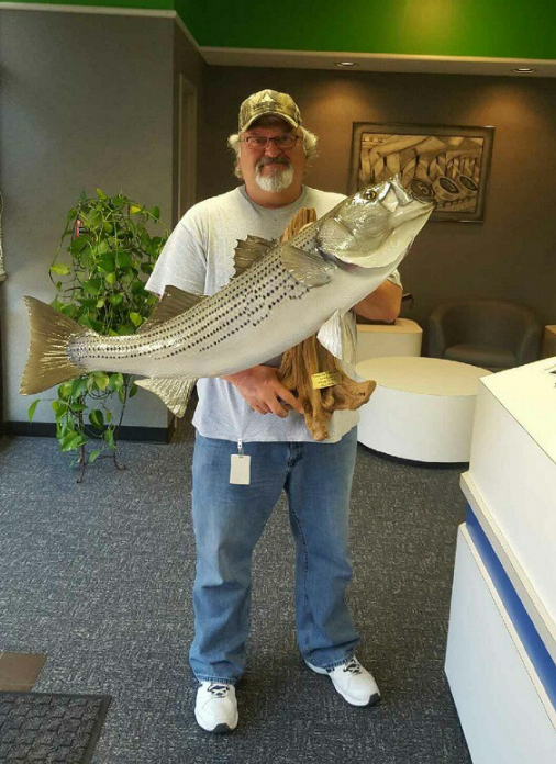 Man holding a large striped bass sculpture indoors, wearing a white shirt and jeans.