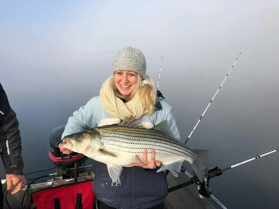 Woman smiling, holding a large striped bass on a boat, foggy lake background.