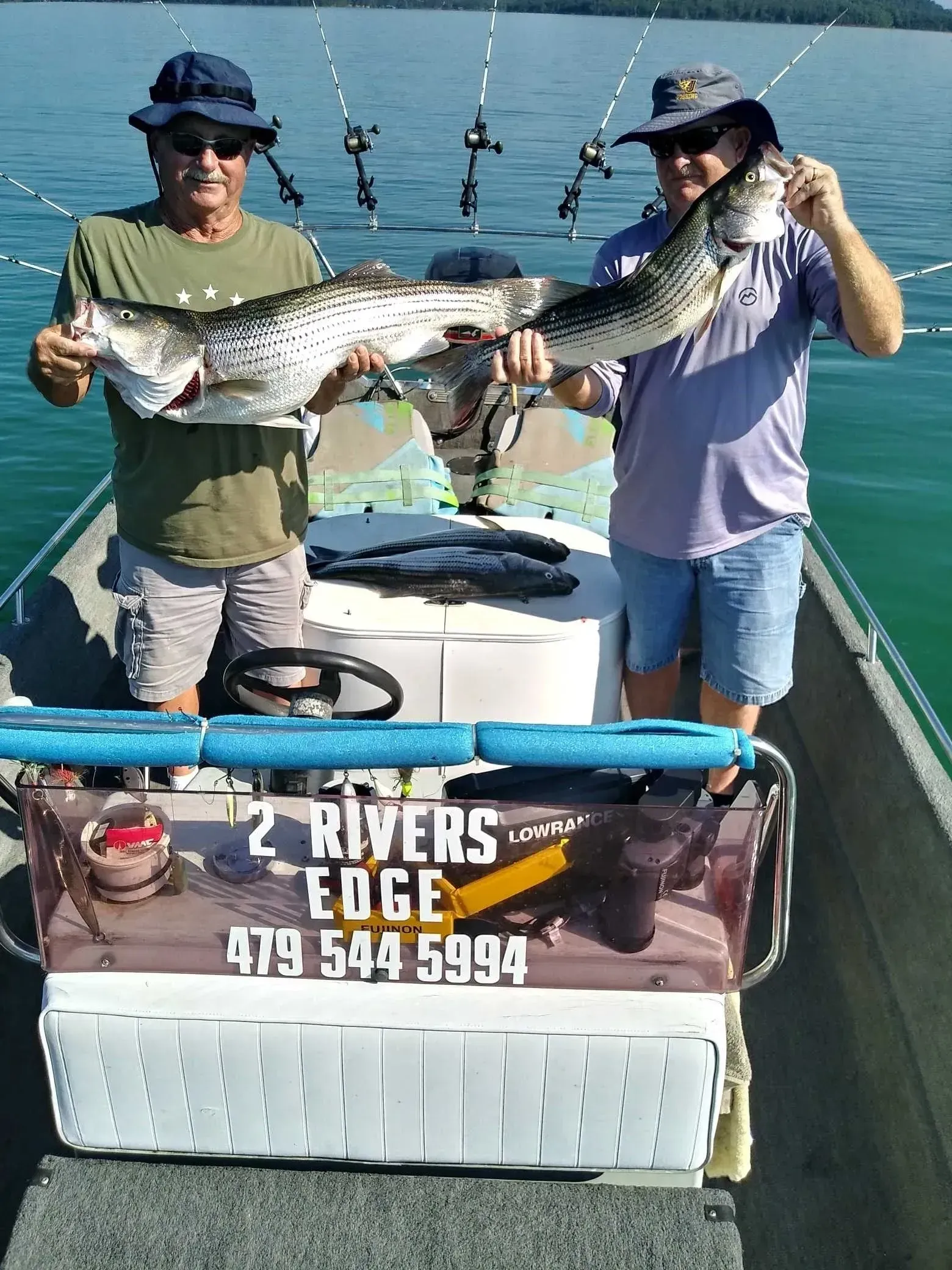 Two men on a boat holding large fish they caught, with fishing rods in the background.