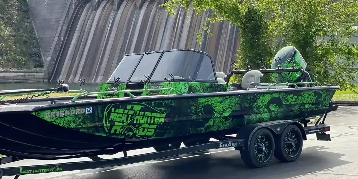 Green and black fishing boat on a trailer parked next to a dam, with green camouflage design.