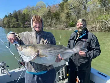 Two people on a boat holding a large striped bass fish. The boy smiles; the other person wears a face covering.