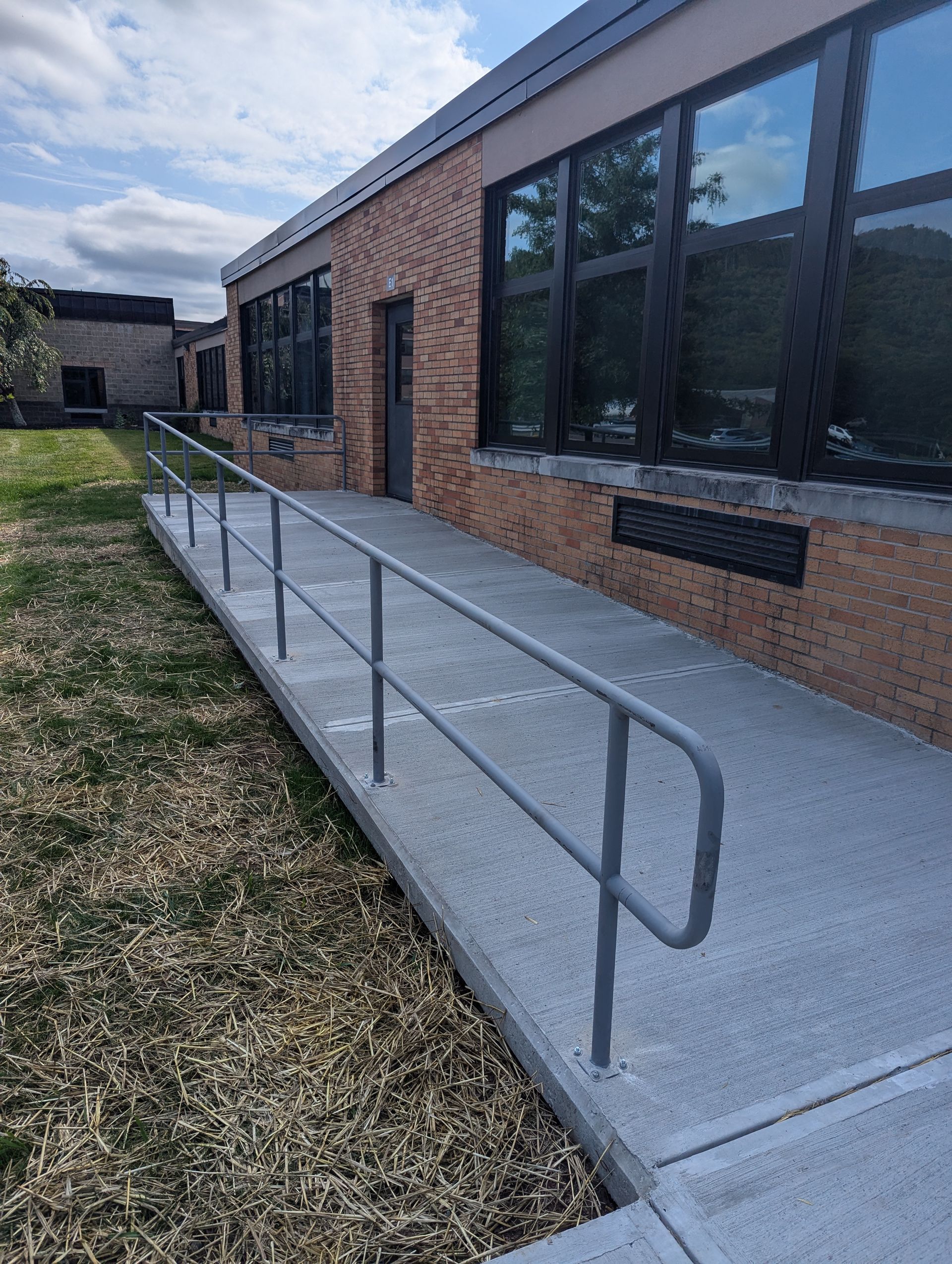 Concrete ramp with gray handrails leading to a building's door. Brick exterior, large windows.