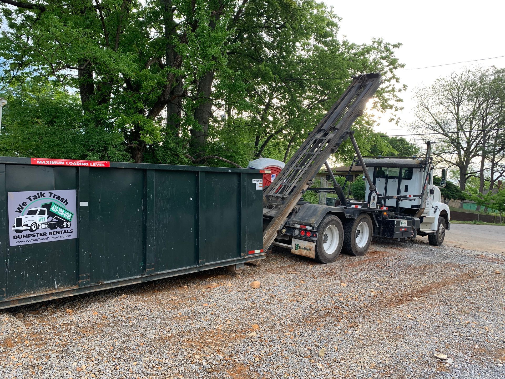 A dumpster is being towed by a dump truck in a gravel lot.