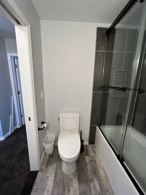 Narrow bathroom with toilet, trash bin, and glass-enclosed shower with gray tile wall.