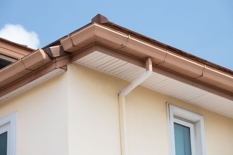 Close-up of a house roof corner with beige siding, brown trim, and a white downspout under a blue sky