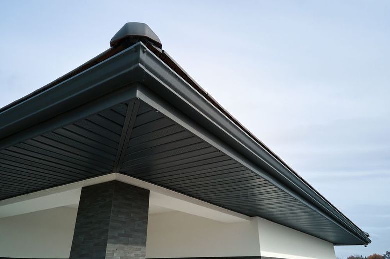 Low-angle view of a house roof with dark trim, gray soffit, and a white wall against a cloudy sky
