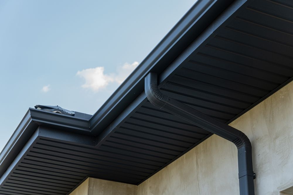 Close-up of a building roof edge with black gutters and downspout against a blue sky.