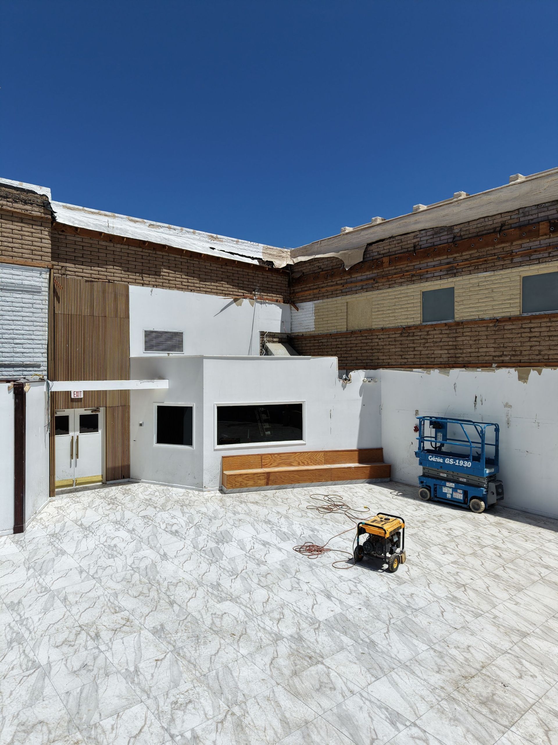 Snow-covered courtyard with a small white building, brick walls, and a blue utility lift under a clear blue sky