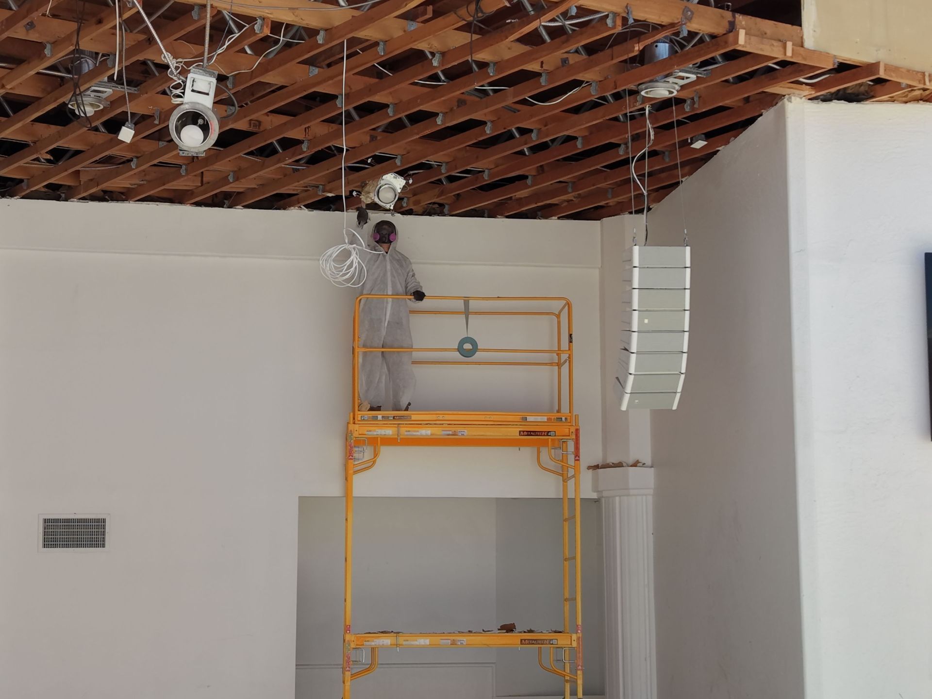 Worker on an orange scaffold installing ceiling fixtures in a room with exposed wooden beams.