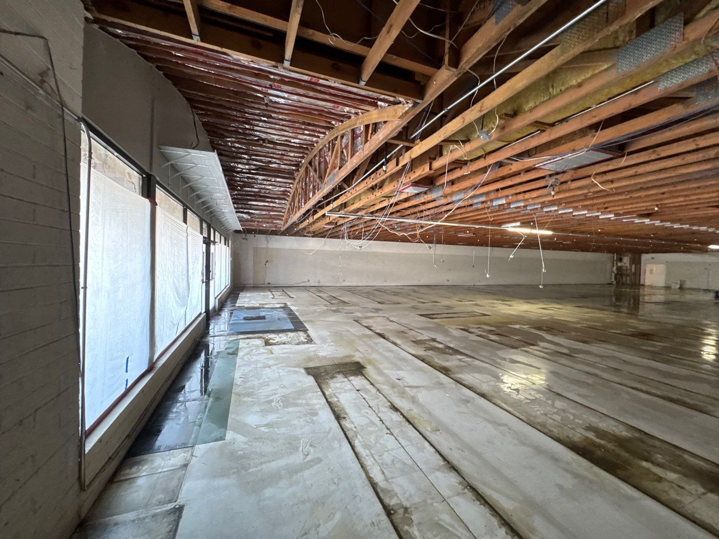 Empty unfinished basement with exposed wooden ceiling beams and concrete floor, lit by windows on the left.