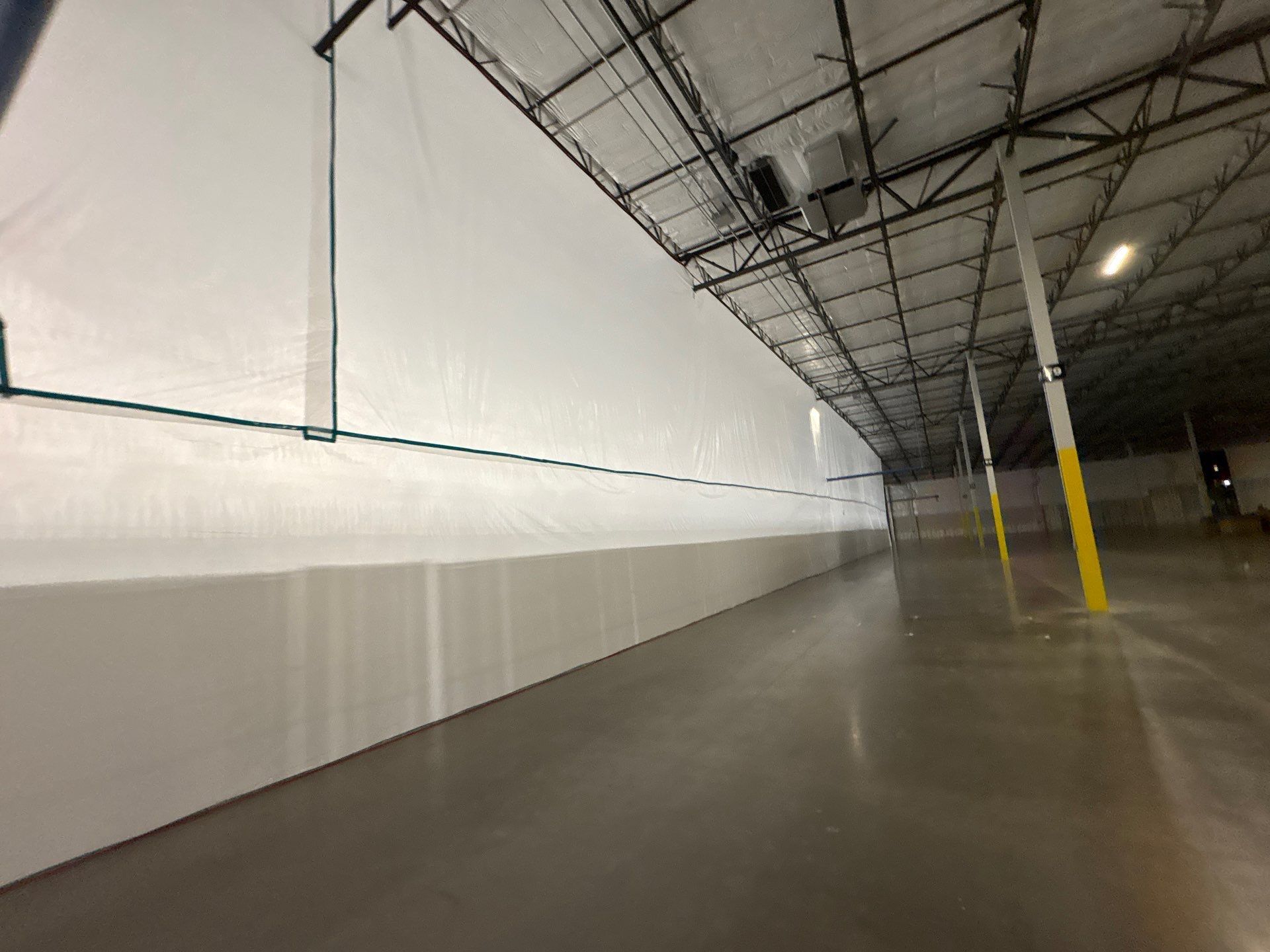 Empty warehouse interior with white wall, concrete floor, and yellow support pillar under exposed ceiling cables