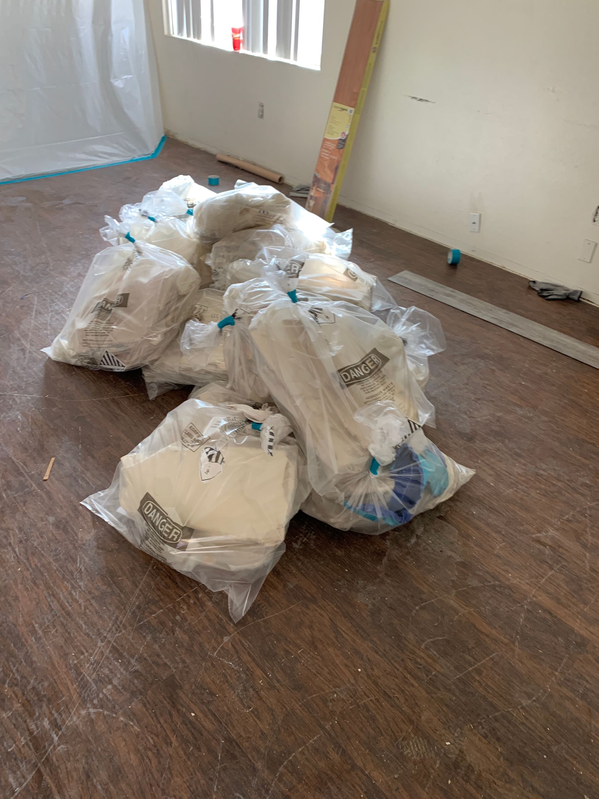 Pile of white trash bags stacked on a hardwood floor in an empty room with a blue tarp and ladder nearby