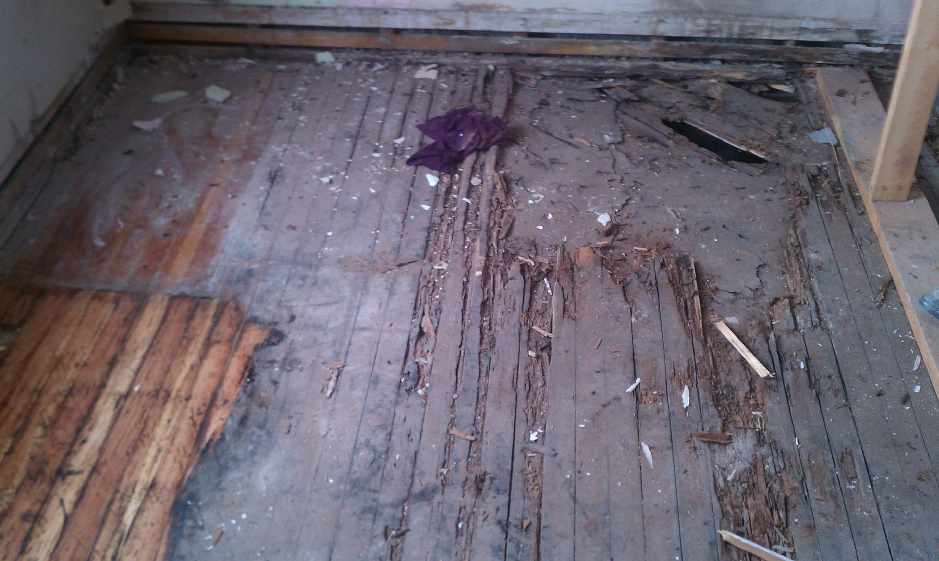 Damaged wooden floor with a dark stain and debris in a corner of a room
