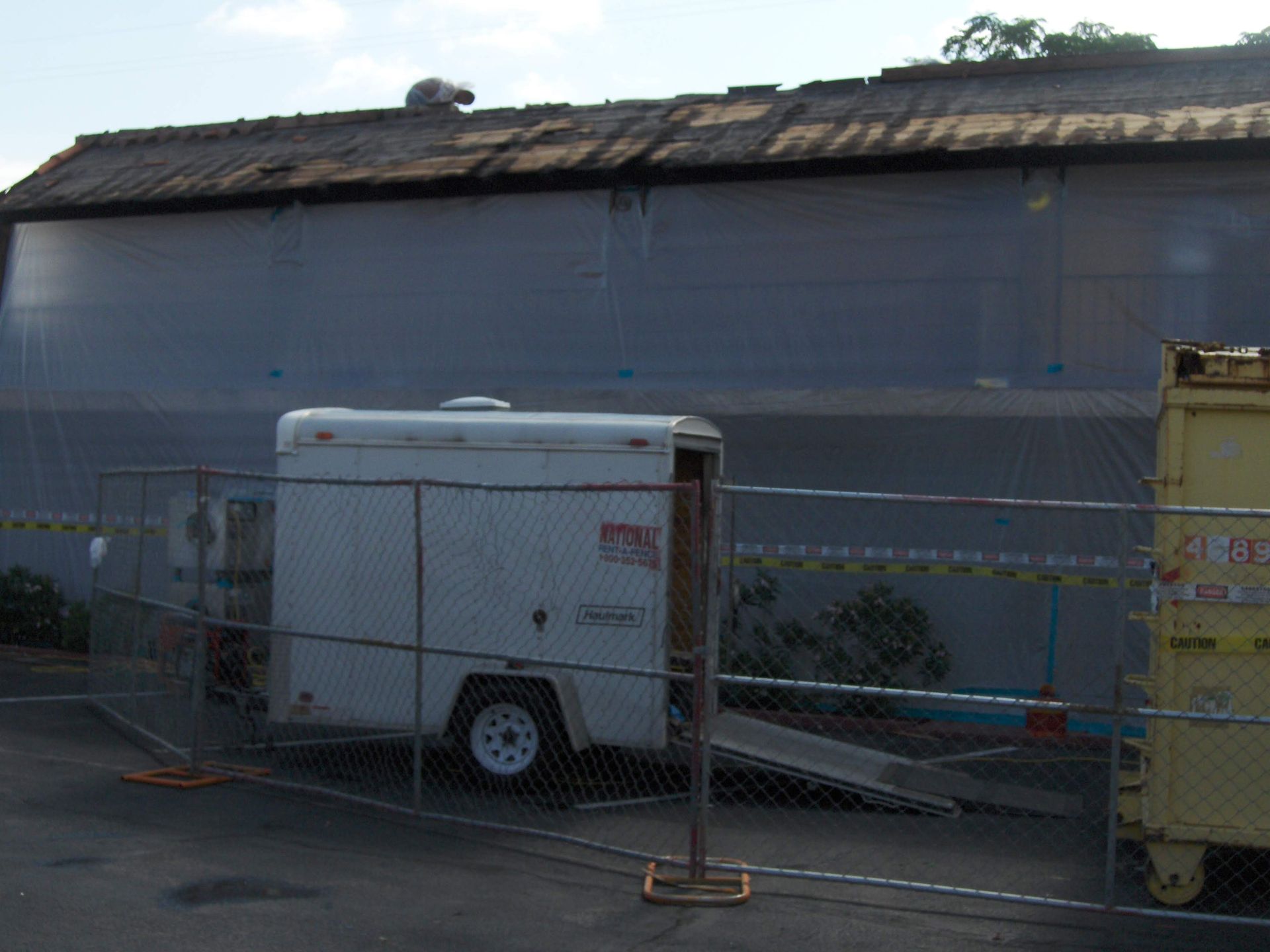 White utility trailer behind chain-link fence beside a building with a yellow dumpster.