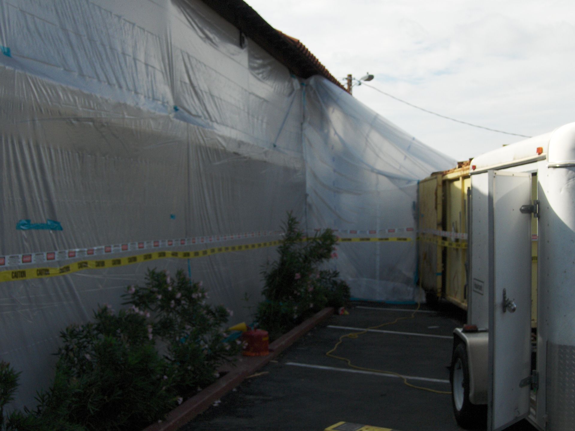 Narrow alley beside a white tarp-covered building, with potted plants and a white trailer.