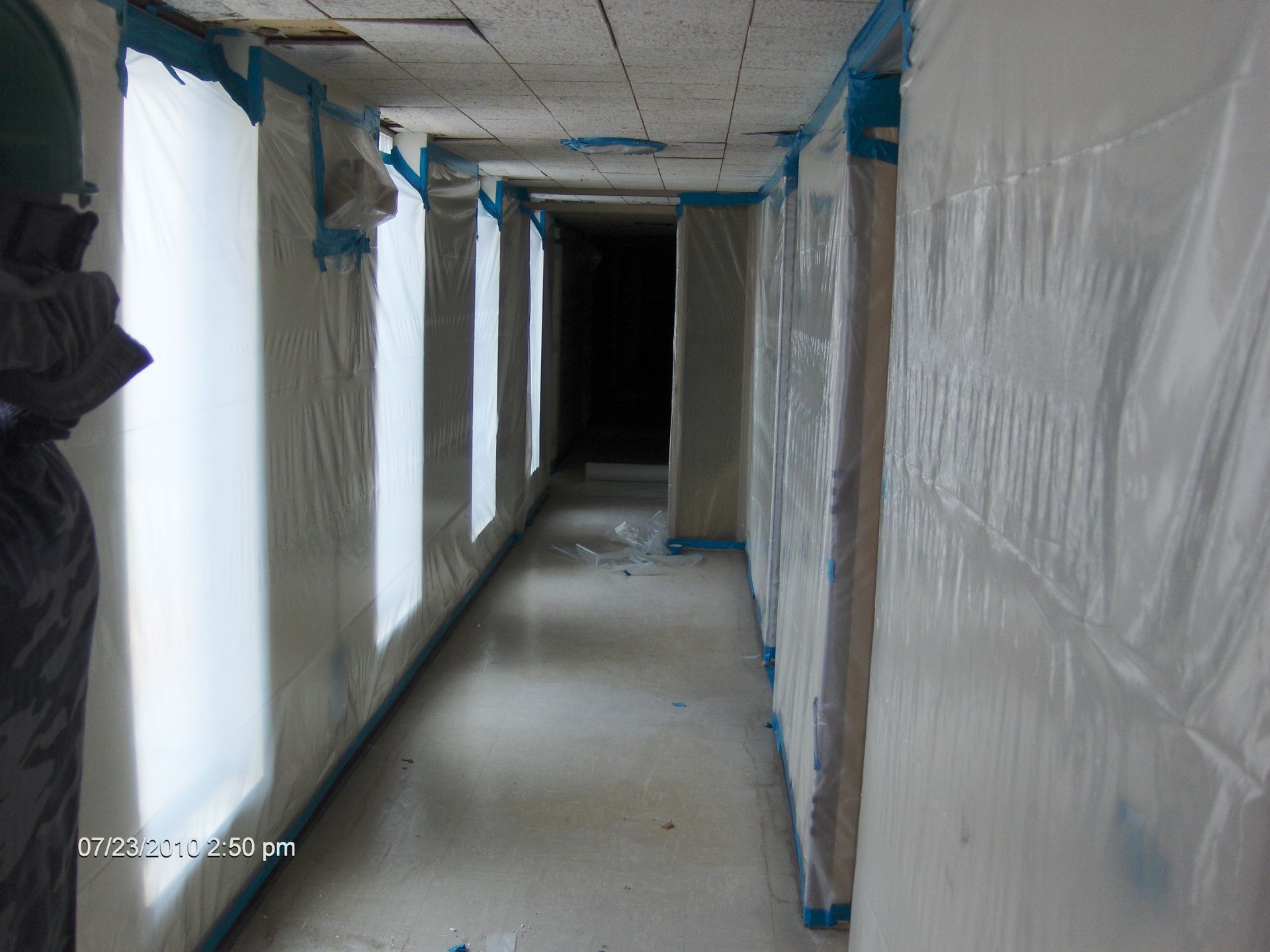 Narrow indoor corridor under renovation, with plastic-covered walls and windows leading to a dark doorway.