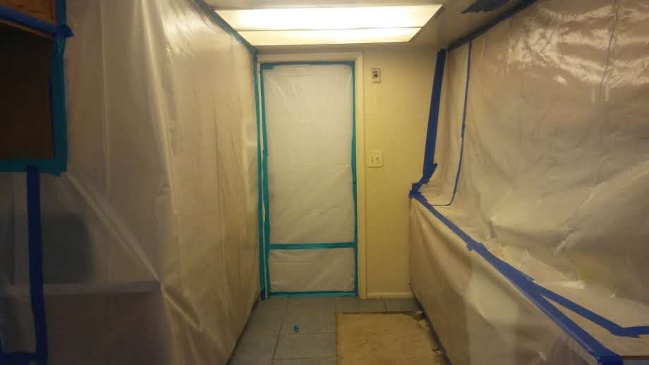 Narrow hallway covered in plastic sheeting, with a taped-off door at the end under bright ceiling lights.