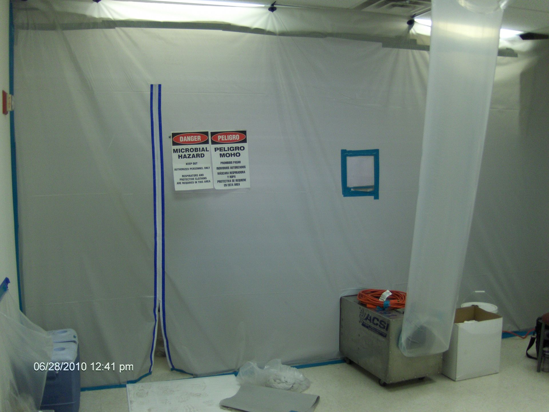 Taped-off room with white plastic walls, warning signs, and a small bin and supplies on the floor