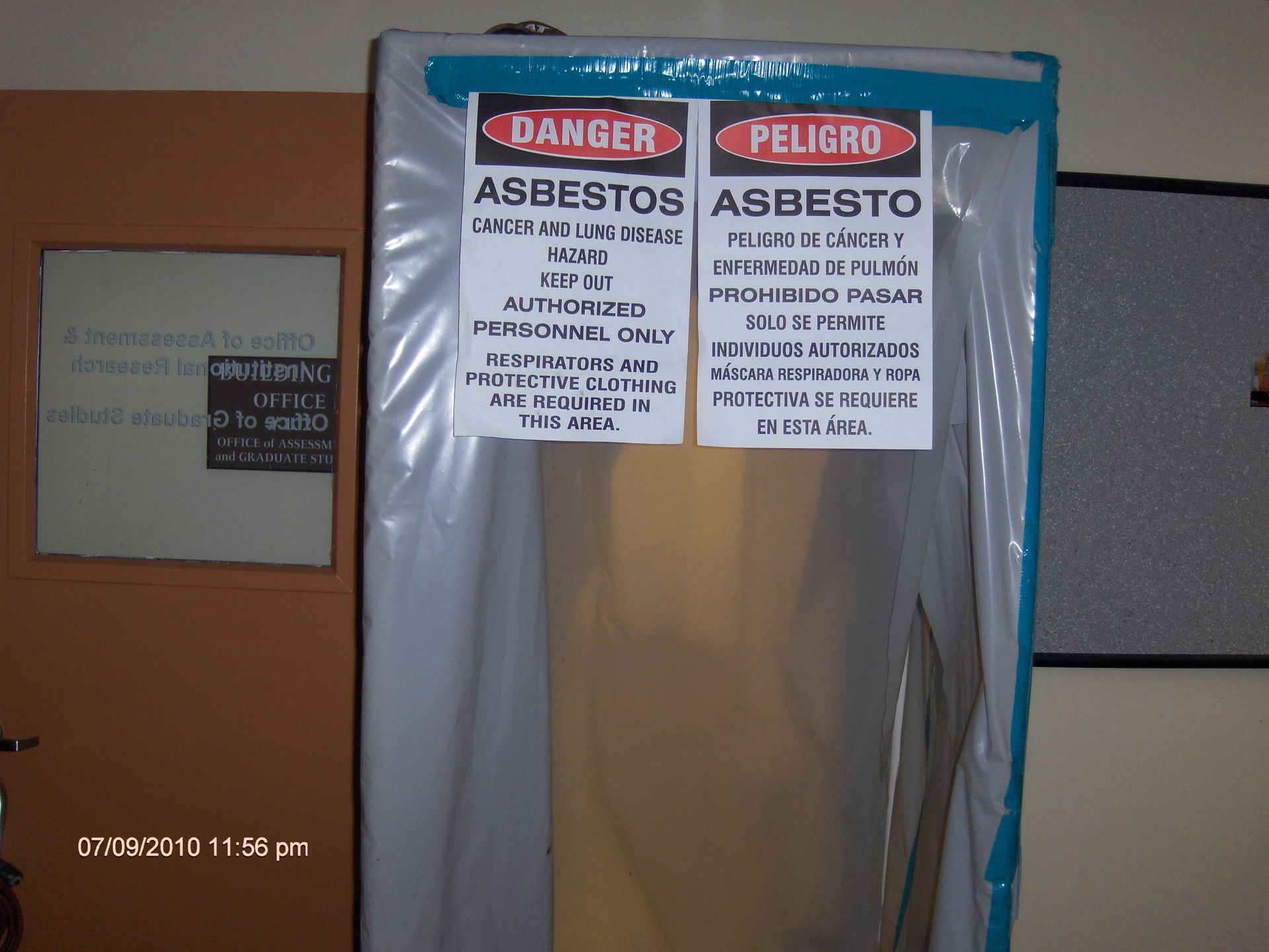Plastic-covered doorway with bilingual asbestos danger signs in English and Spanish.