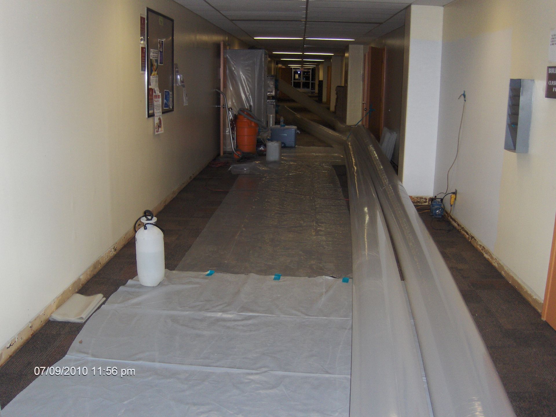 Narrow hallway under renovation with plastic sheeting, hoses, and construction materials on the floor.