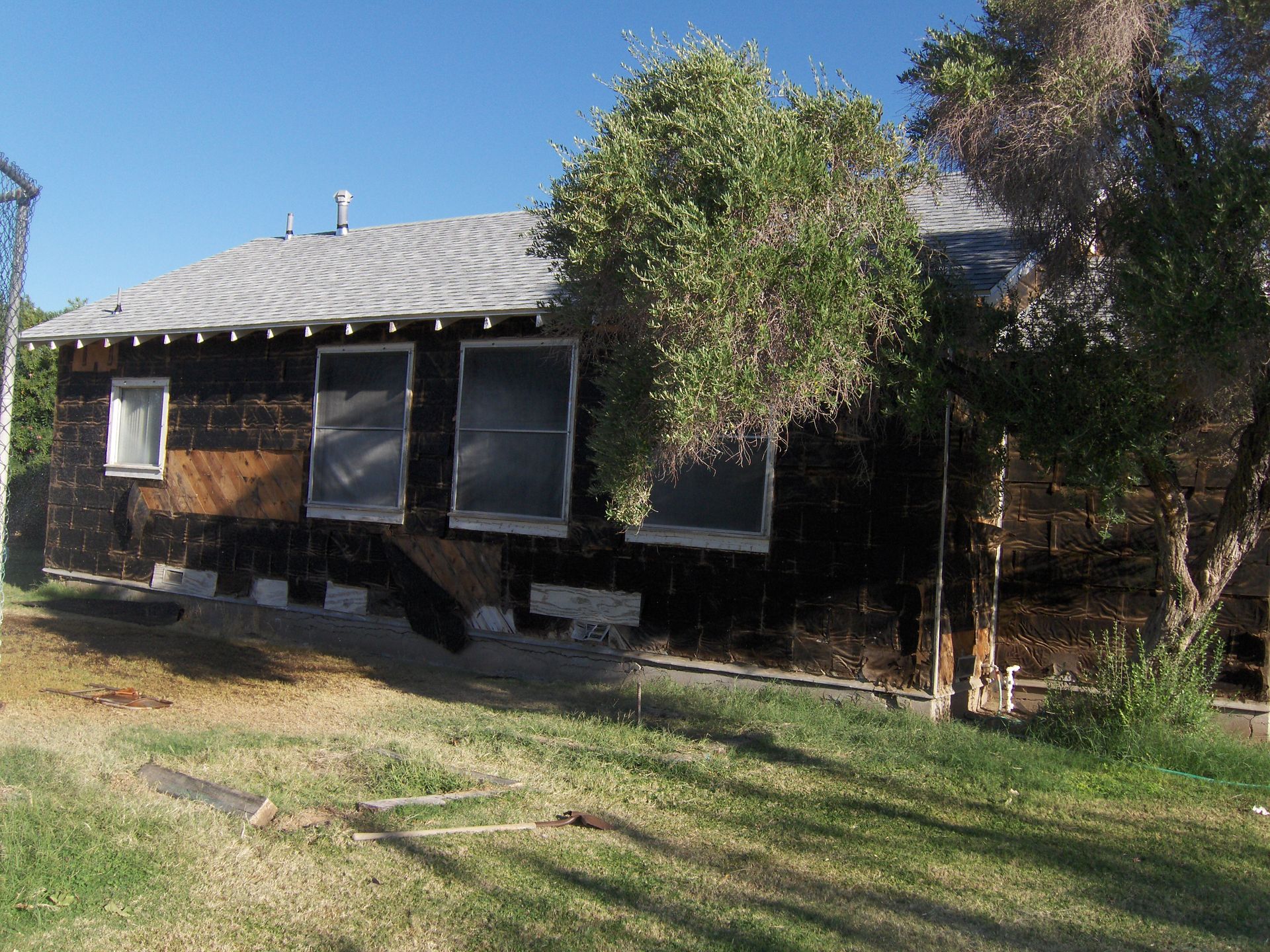 Small dark house with a light roof, front porch, and trees in a sunny yard.