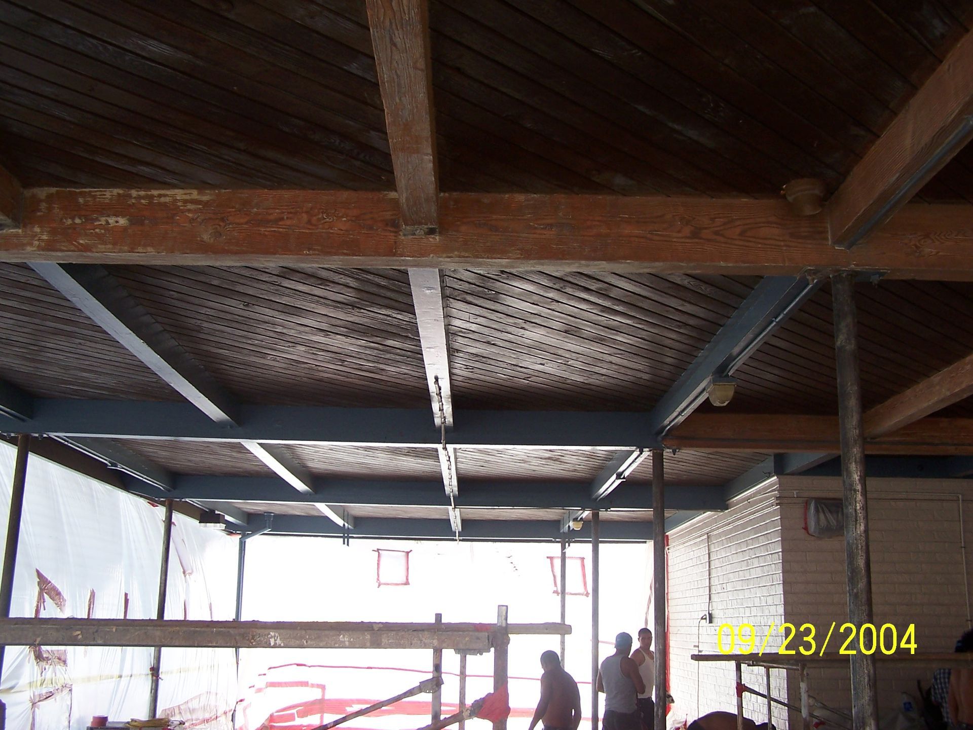 Interior with exposed wooden roof beams, a brick wall, and people standing near scaffolding in a work area