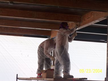 Two workers in white coveralls installing a ceiling beam beneath a wooden structure.