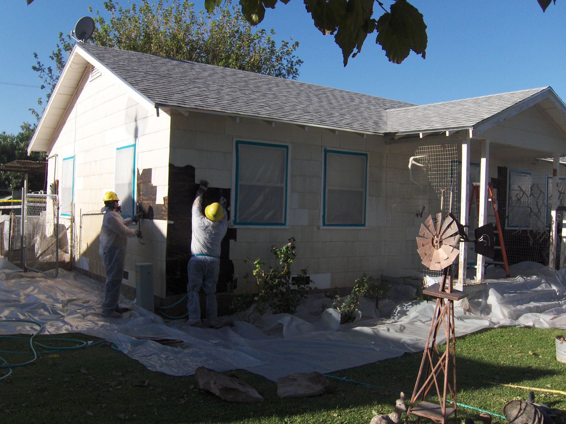 Workers applying stucco to a small house exterior with scaffolding and tools in the yard