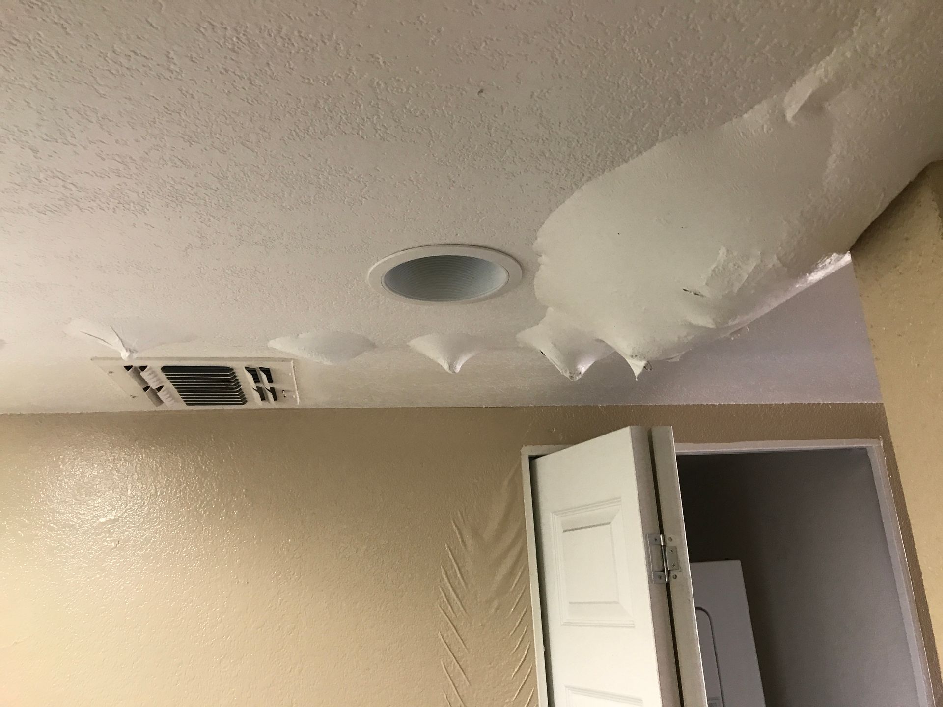 Damaged ceiling with peeling drywall and a circular vent above an open white door in a beige room
