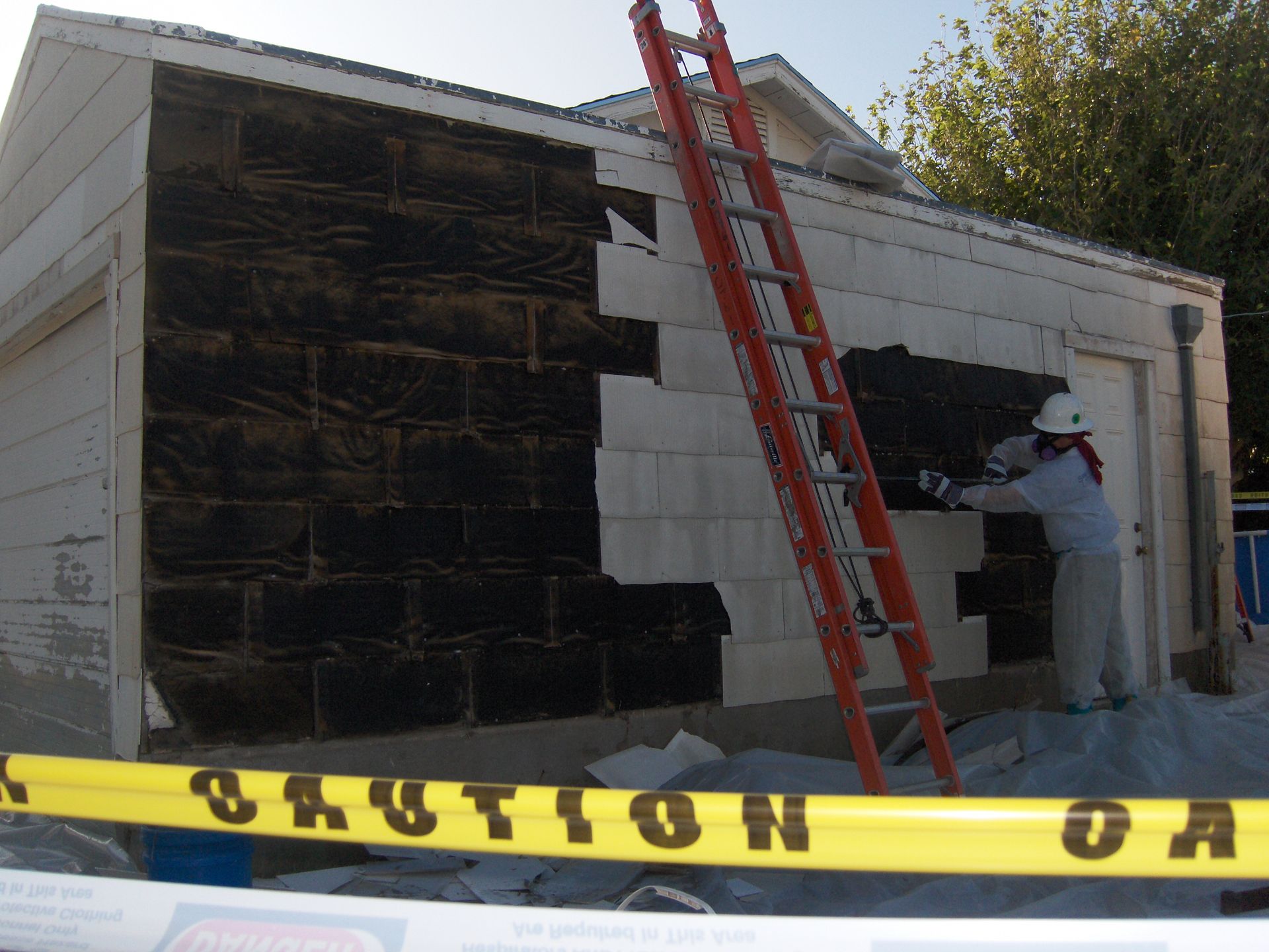 Two workers repairing a white wall, with a red ladder and black tarp, behind yellow caution tape.