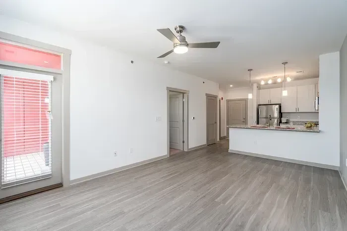 Empty modern apartment living area with gray floors, white walls, ceiling fan, and open kitchen.