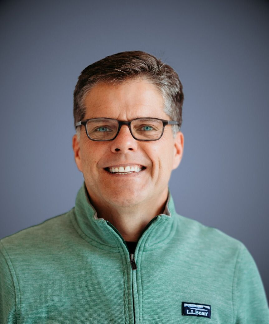 Man wearing glasses and a green zip-up sweater smiles at the camera against a blue background.