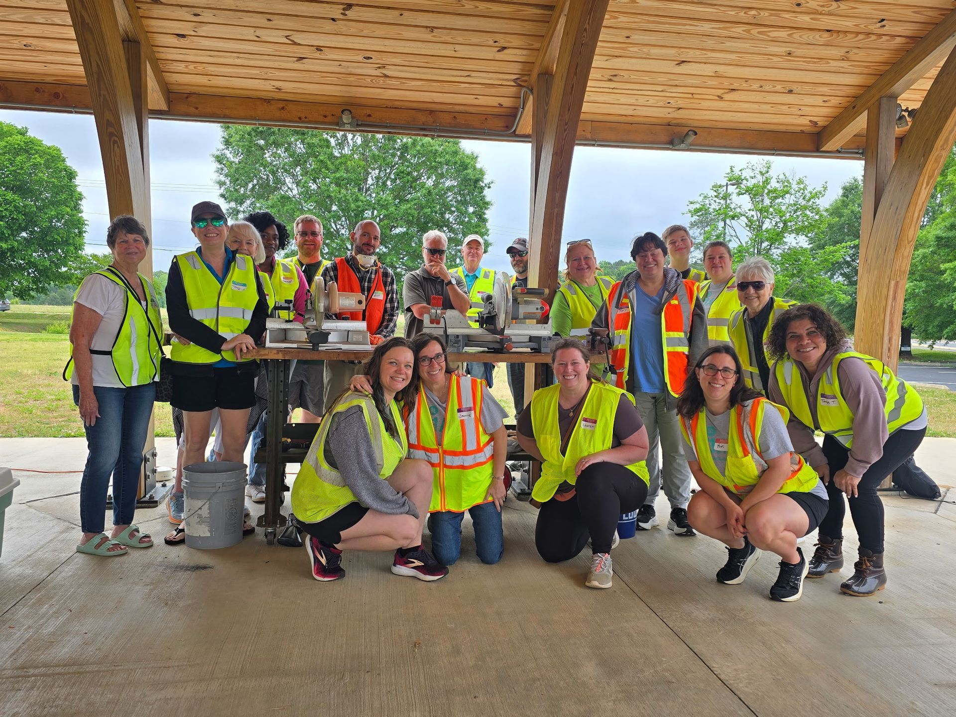 Group of people wearing yellow vests, smiling and posing under a wooden shelter outdoors.