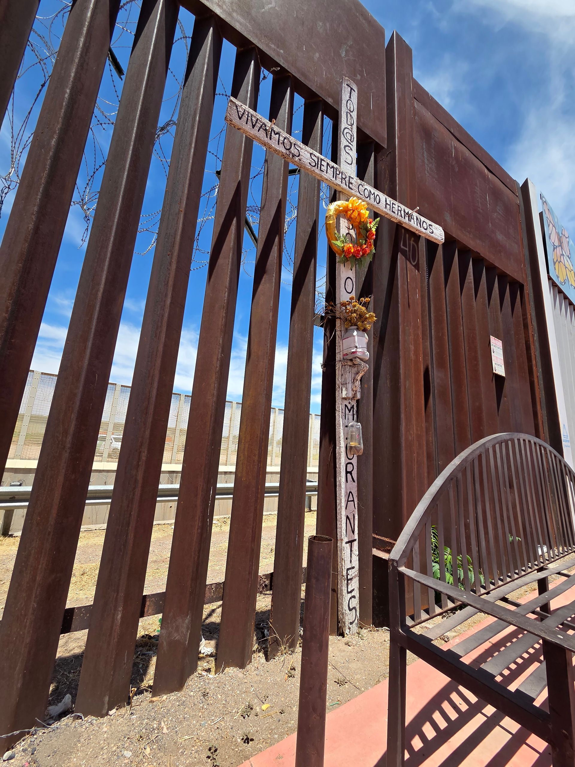 A cross memorial with flowers against the U.S.-Mexico border wall.