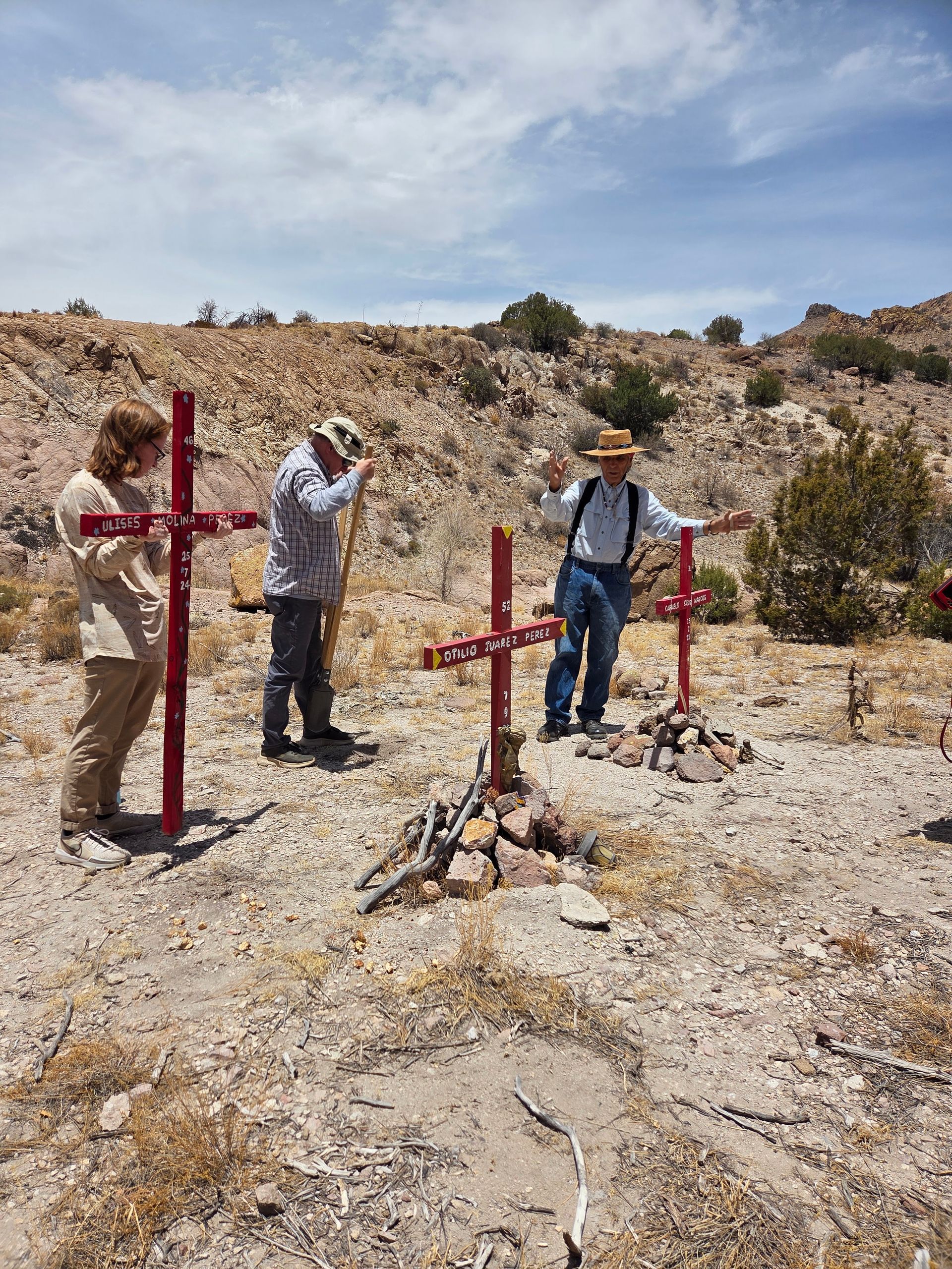Three people stand by red crosses in a desert, arms outstretched, under a sunny sky.