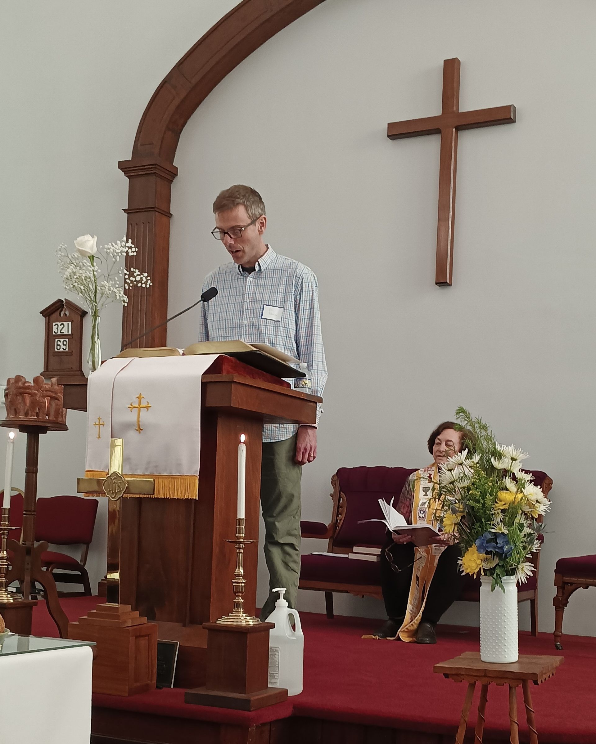 A man reads from a podium in a church; a woman sits nearby with flowers; a wooden cross is on the wall.