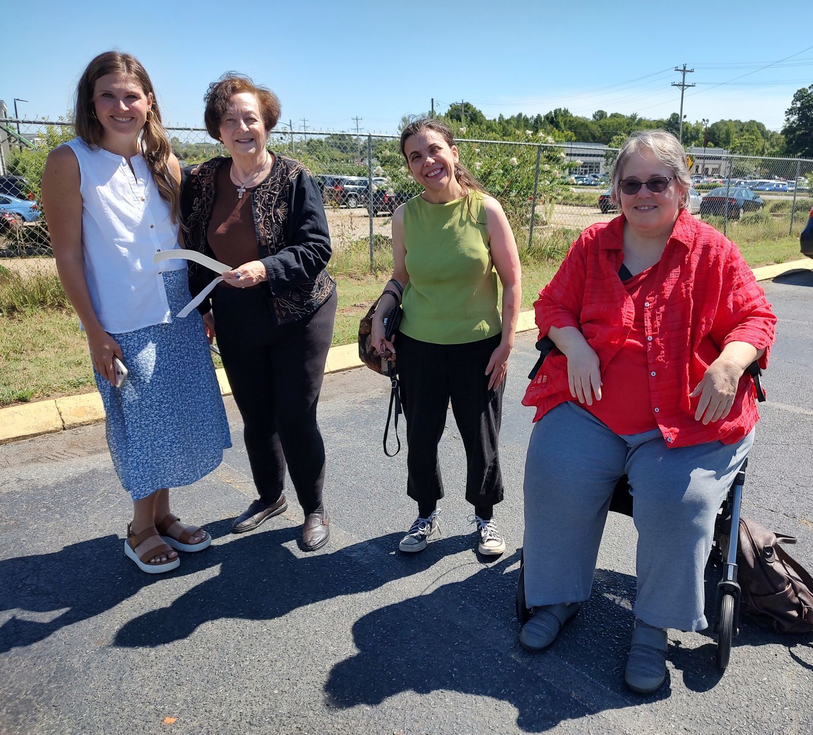 Four women stand and sit outside on a sunny day. One woman is in a wheelchair and wears a red shirt.