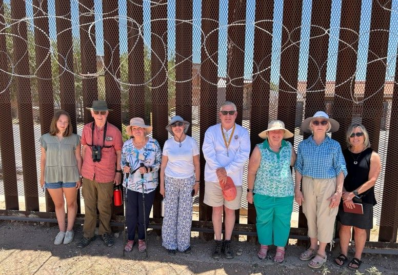 Group of people standing in front of a tall border fence with barbed wire in sunny outdoor setting.