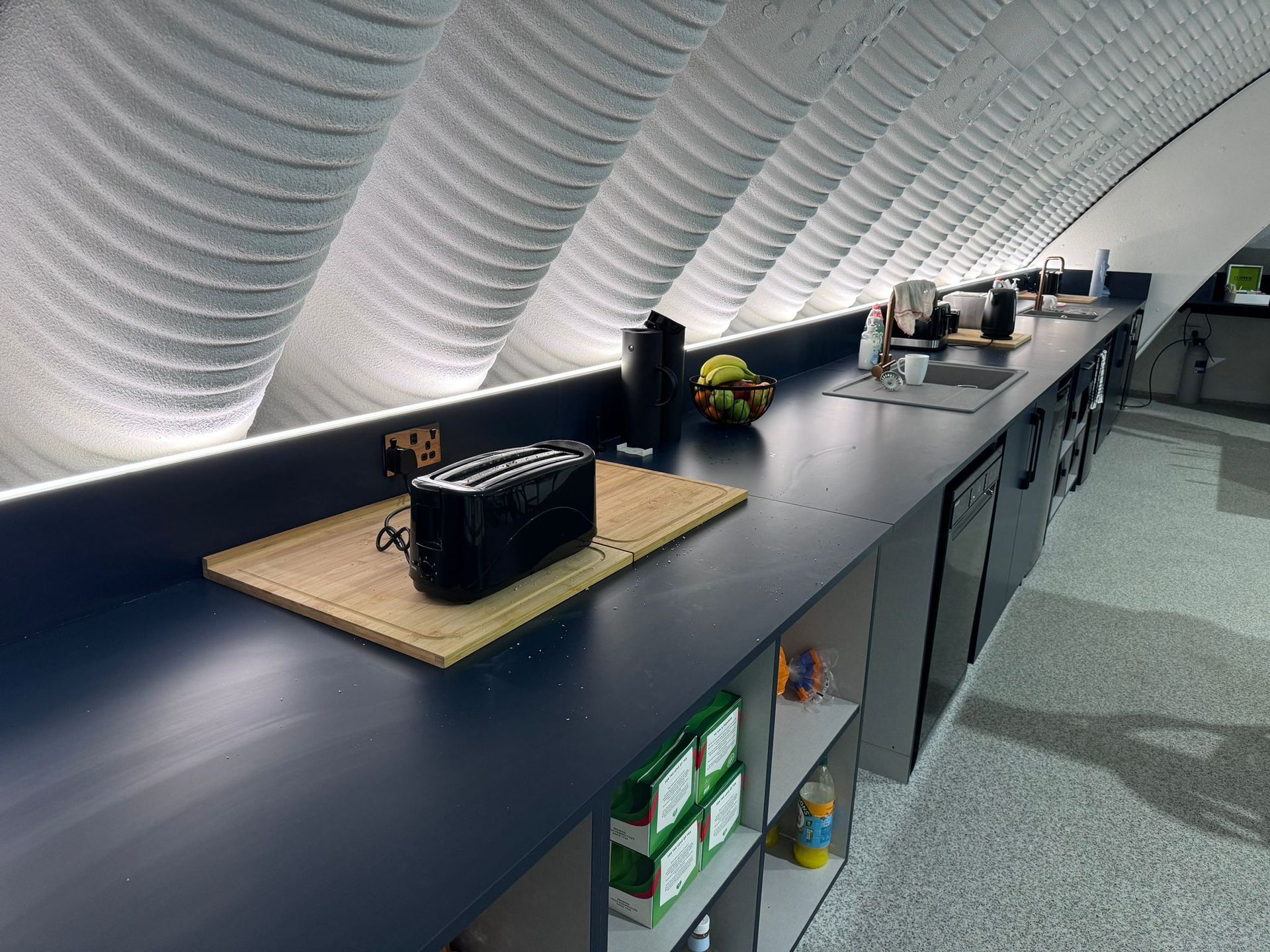 A modern kitchen counter with a black toaster, a bowl of fruit, and a sink, set beneath a uniquely sloped, ribbed ceiling.